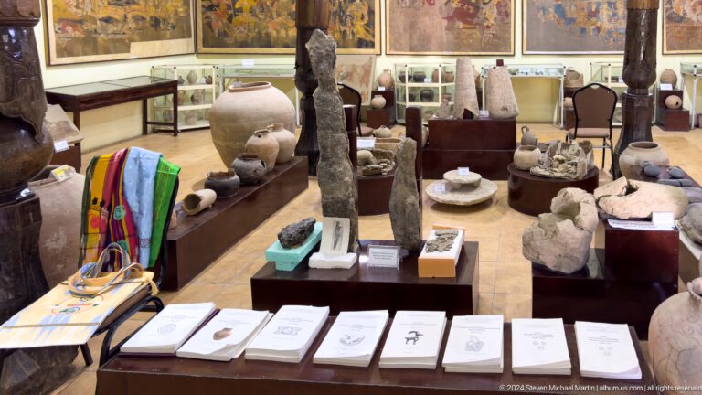Study-collection room at a Panjakent museum with shelves and cases holding pottery and stone artifacts, and wall panels displaying mural reproductions.
