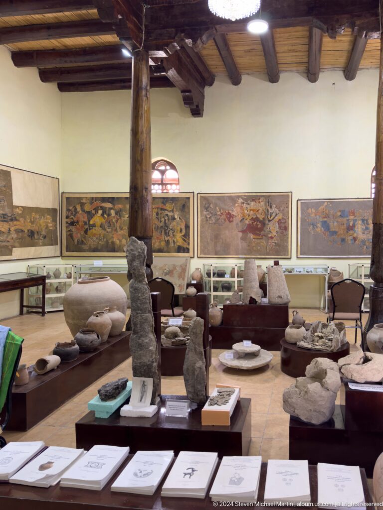 Wide interior view of a Panjakent museum gallery with Sogdian wall painting fragments or reproductions mounted above display cases of archaeological artifacts and wooden columns in the room.