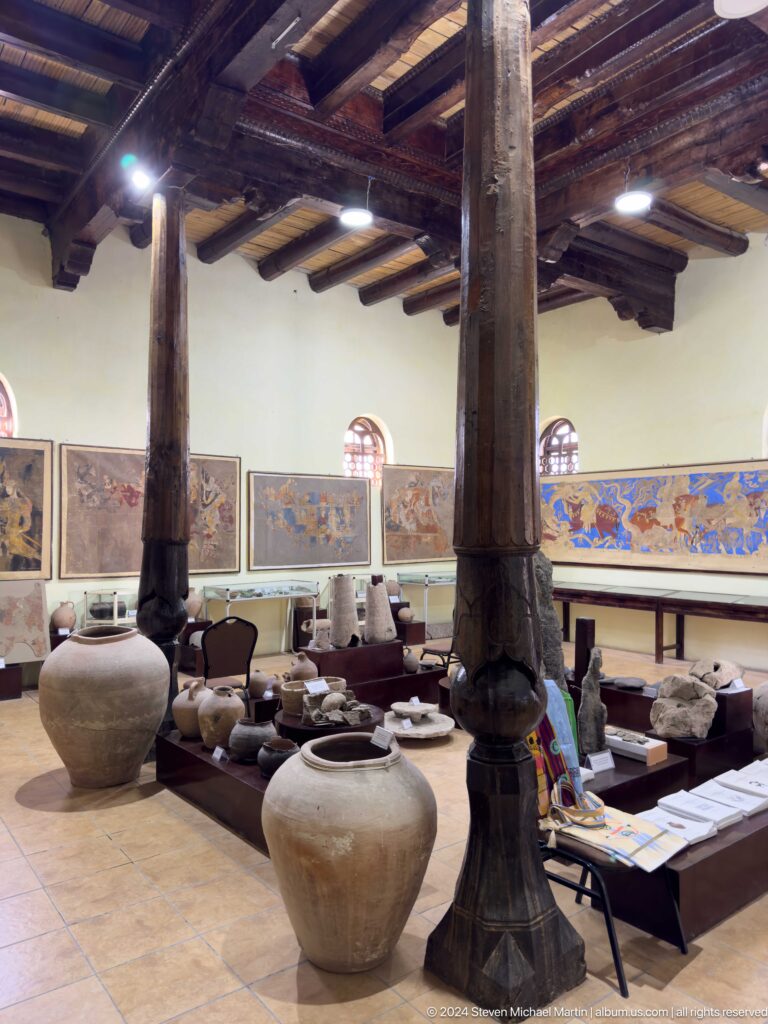 Interior of a Panjakent museum gallery with wooden columns, large pottery jars on the floor, and reconstructed Sogdian wall paintings running around the walls.
