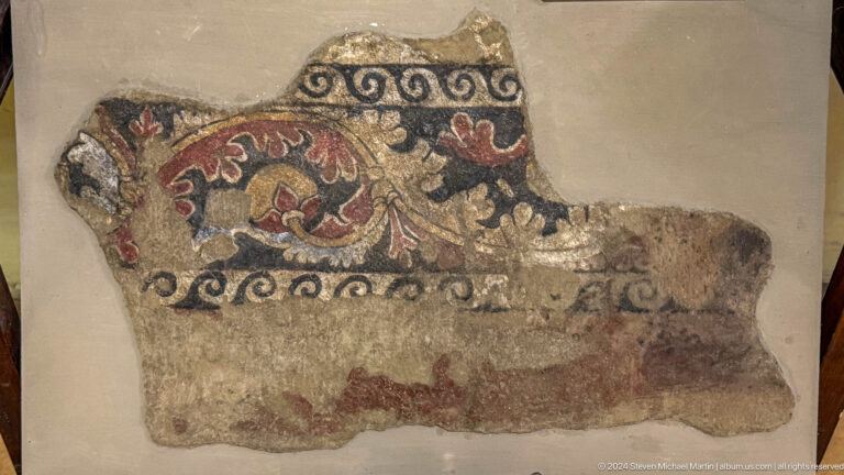 Close view of a painted wall painting fragment from Ancient Panjakent with scrolling vegetal ornament and a black wave border on plaster.