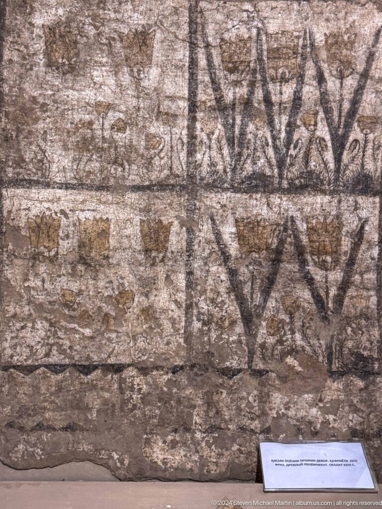 Close view of a worn wall painting fragment from ancient Panjakent, showing faded horizontal bands of dark repeating geometric or vegetal motifs on plaster.