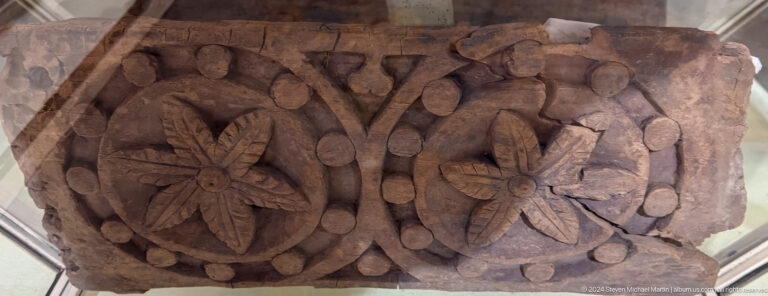 Close view of a dark wooden relief panel from Panjakent, carved with symmetrical floral and leaf patterns around a central palmette.