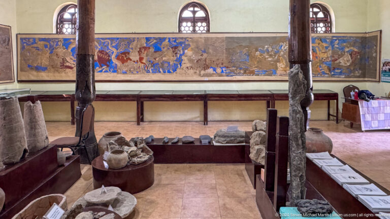 Museum gallery with a long glass case displaying restored Panjakent wall paintings, flanked by timber columns and cases holding excavated artifacts.