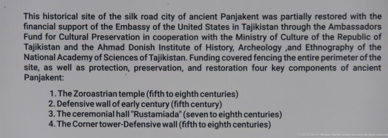 Detail of an information panel for the Ancient Panjakent preservation project, showing English text listing conserved structures such as a Zoroastrian temple, shrines, defensive walls, and gates.