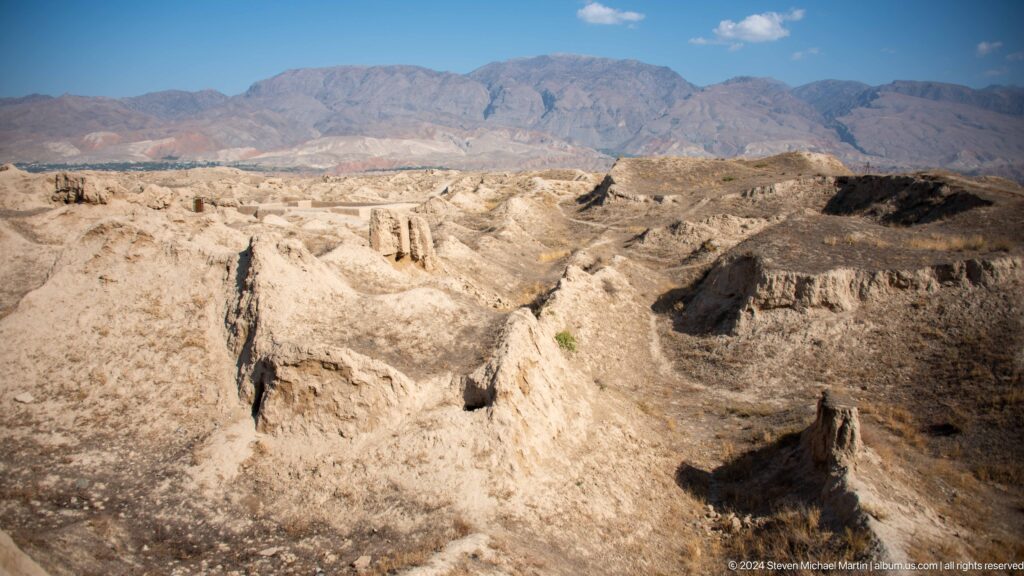Panoramic elevated view of low earthen and mud-brick ruins at Ancient Panjakent spread over a dissected hilltop, with mountains in the background.