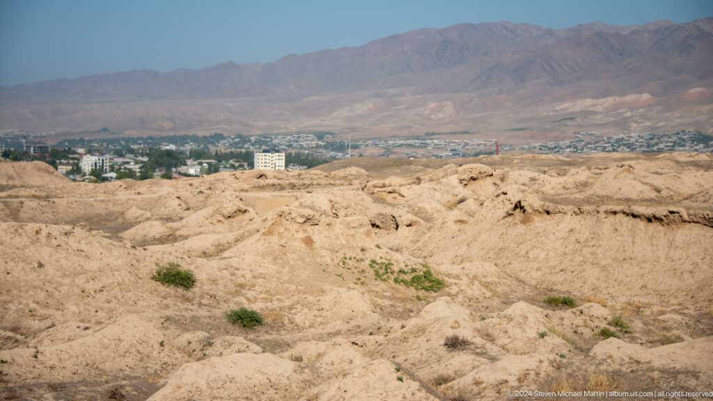 Wide view from the dry hilltop ruins of ancient Panjakent overlooking the greener modern city in the valley with mountains in the distance.
