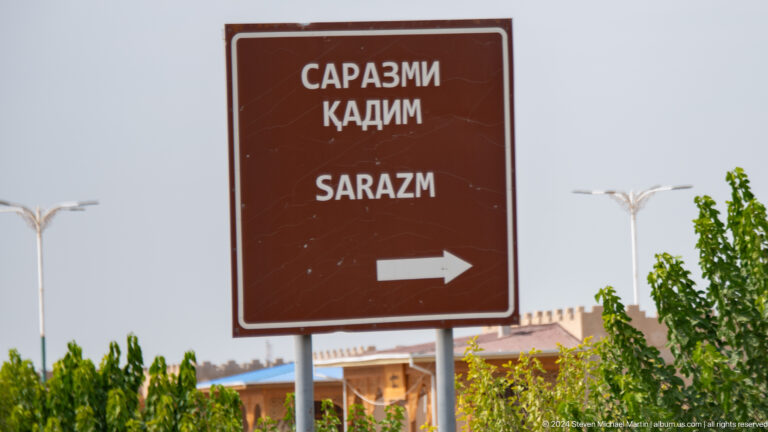 Brown trilingual road sign reading 'Sarazm' with a right-pointing arrow, in Tajik, Russian, and English, along a roadside.