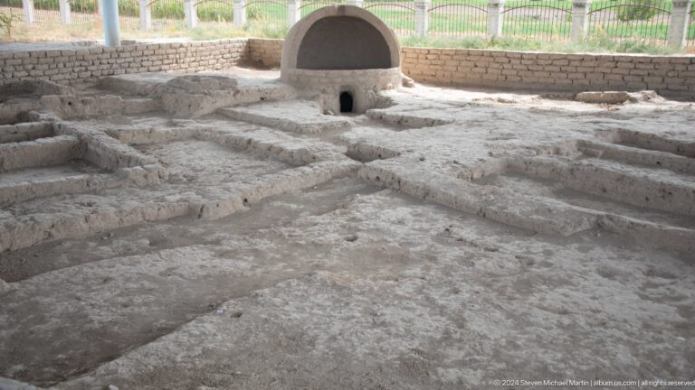 Excavated mud-brick rooms at Ancient Sarazm with low partition walls and a domed earthen installation at the back under a protective shelter.