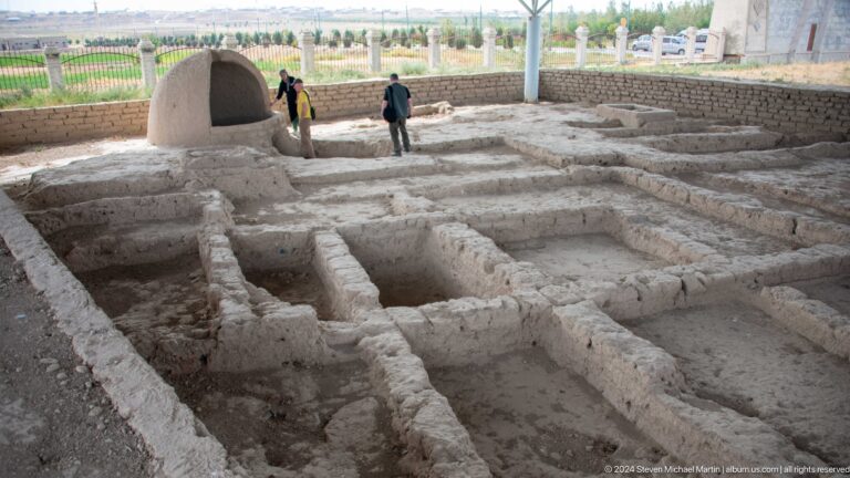 Roofed shelter covering low mud-brick walls of an archaeological excavation, with a person standing among the walls.