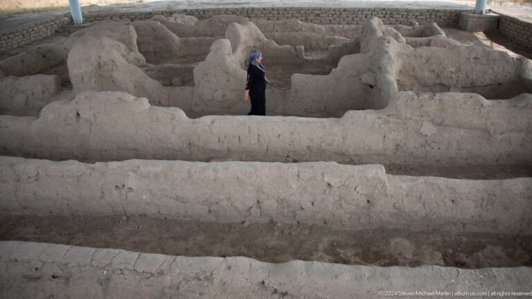 Visitor walking between low, parallel mud-brick walls of an excavated room and corridor at Ancient Sarazm, beneath a protective shelter.