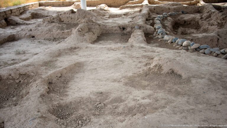 Shallow excavated area at Sarazm with an irregular compacted floor, low walls, and a circular depression.