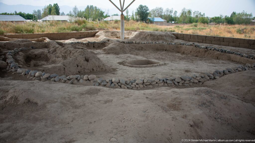 Wide view of a large rounded excavation at Sarazm, enclosed by a curved boundary with stones along part of the edge.