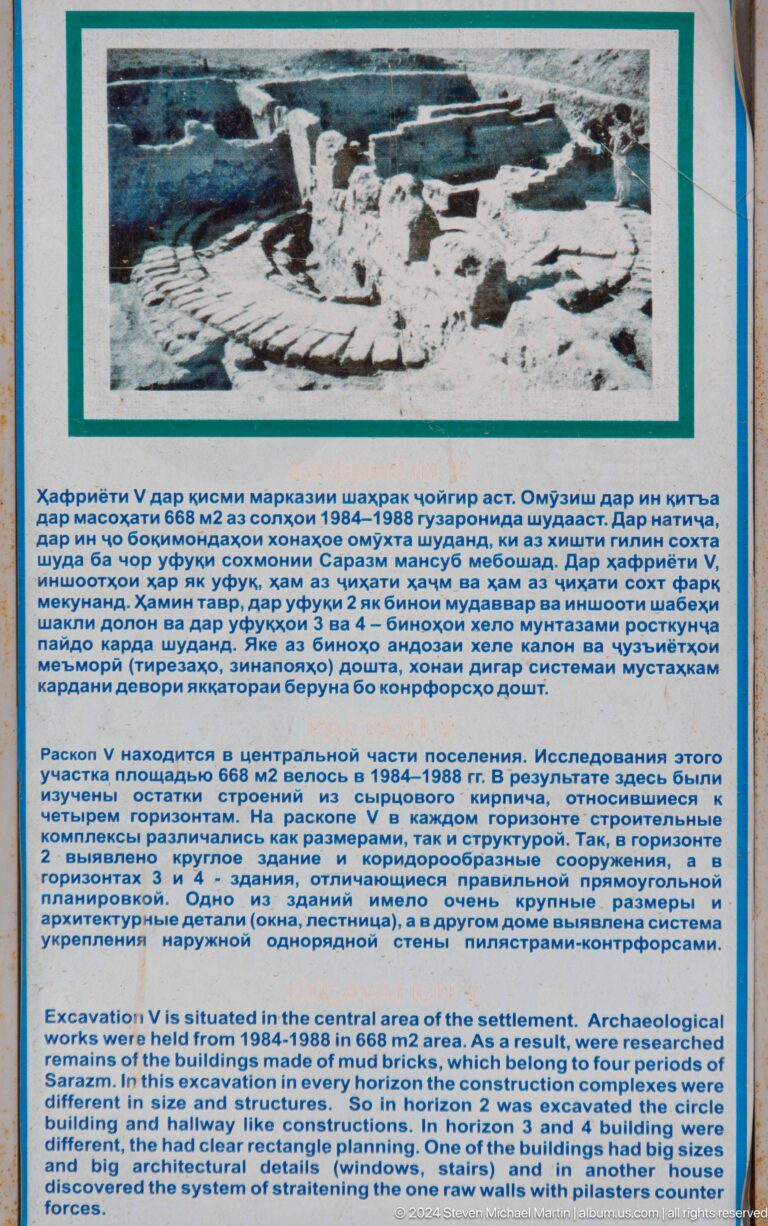 Close view of a blue-bordered interpretive panel at Sarazm with Tajik, Russian, and English text and a small illustration above the main text.