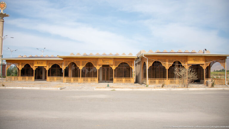 Long open-fronted visitor structure with repeated arches at Ancient Sarazm, standing beside an open paved area.