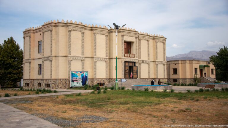 Wide frontal view of the pale, symmetrical Ancient Sarazm museum or visitor center with interpretive signs on the open grounds in front.