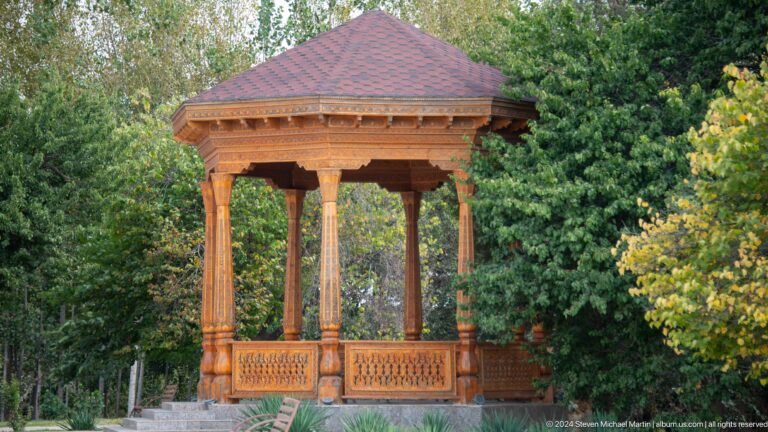 Ornately carved wooden pavilion at the Sarazm museum, with a hipped roof and carved columns around an open seating platform among dense trees.