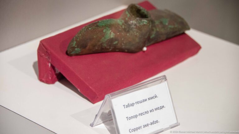 Small irregular elongated artifact from Sarazm displayed on a red platform with a museum label.