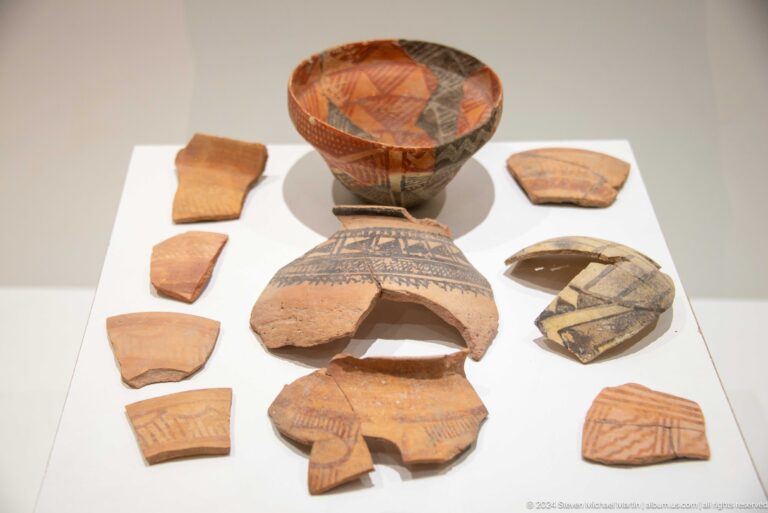 Painted and incised pottery from Sarazm, featuring a red-painted vessel behind linear-decorated sherds in a museum display case.