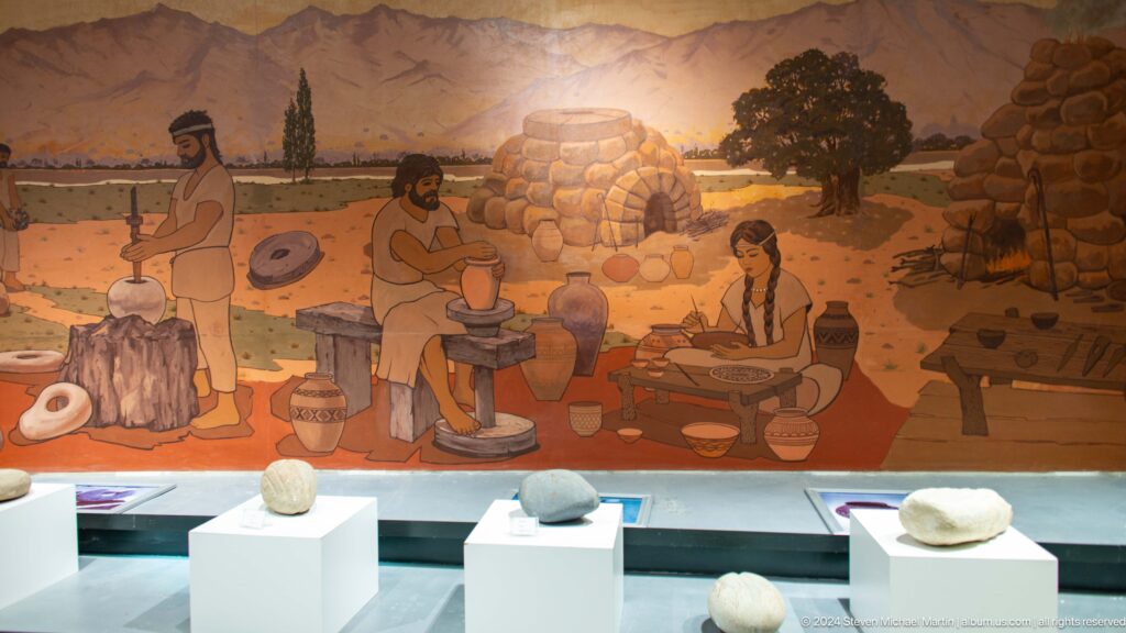 Museum mural reconstructing daily life at ancient Sarazm, with figures farming, crafting, and tending animals above artifact display cases.