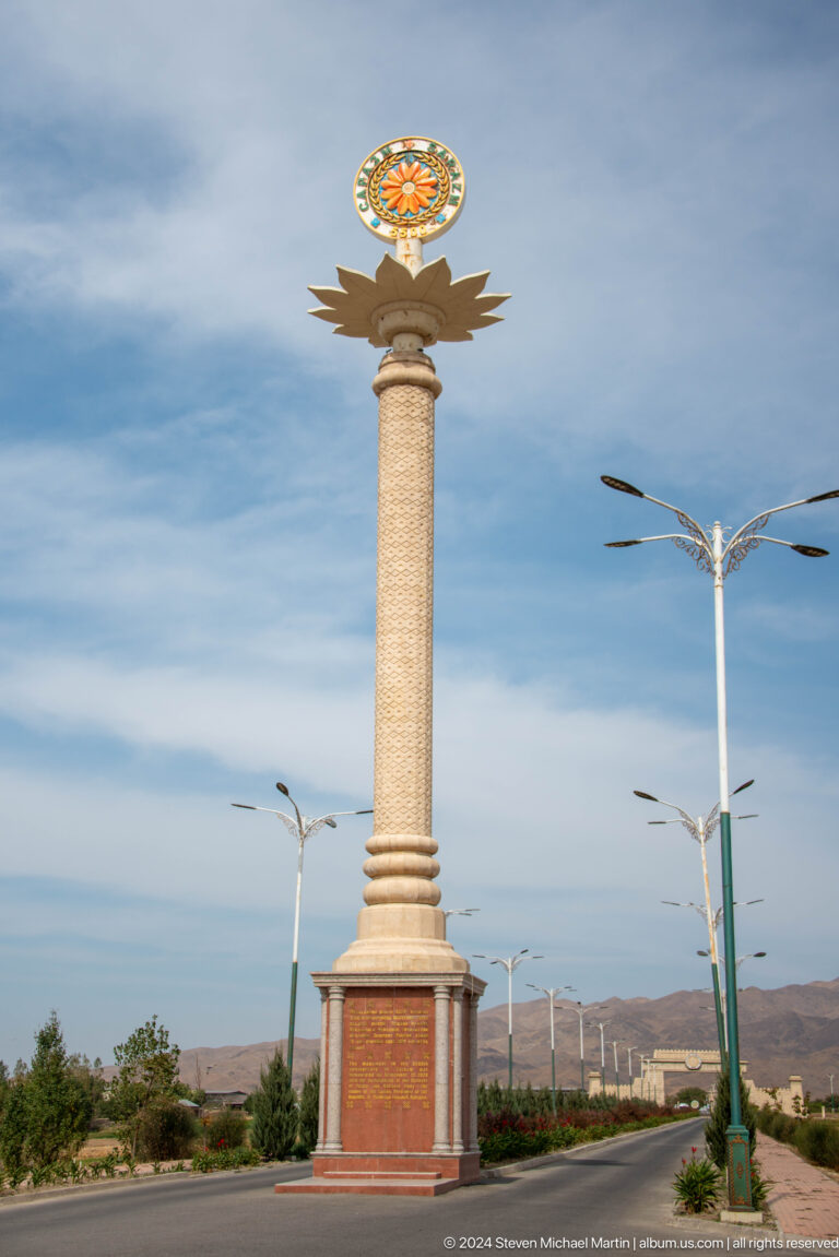 Tall commemorative column with an ornamental capital at the Sarazm entrance, surrounded by modern streetlights under a blue sky.