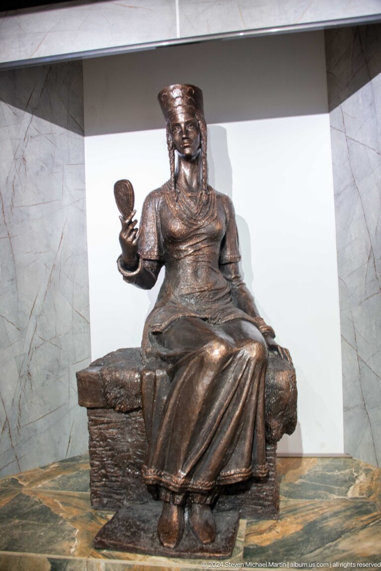 Modern bronze sculpture of a seated female figure in a long garment, one hand raised holding a small object, associated with Sarazm and displayed in a museum.