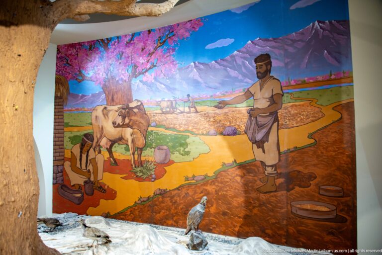 Colorful museum mural showing prehistoric figures farming and tending domestic animals in a mountainous, cultivated landscape at ancient Sarazm.