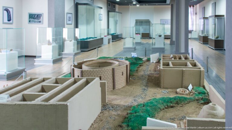 Archaeological exhibition gallery at Sarazm with low white display cases and a large central architectural model.