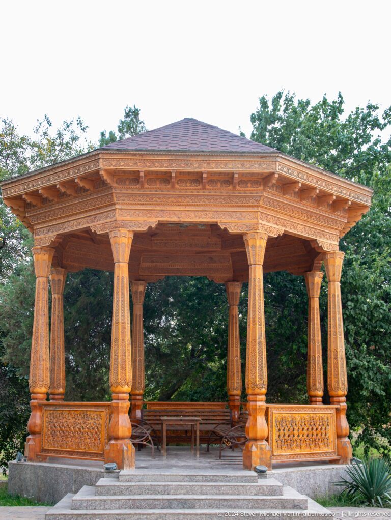 Close view of a carved wooden pavilion with geometric and vegetal patterns on beams and columns, supported by multiple slender posts at the Sarazm complex.