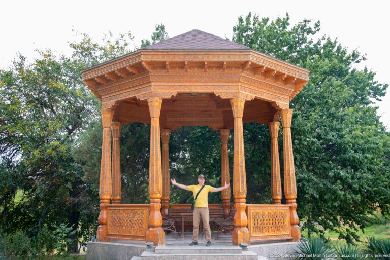 Full view of an open octagonal carved wooden pavilion in the Sarazm museum grounds, with a seated visitor inside for scale.