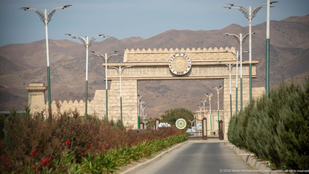 Approach road leading through the ceremonial entrance gate to Ancient Sarazm, with a circular Sarazm emblem above the opening.