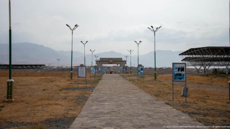 Straight paved walkway through the Sarazm archaeological reserve with interpretive signs and shelters on dry open ground, leading toward a distant low building.