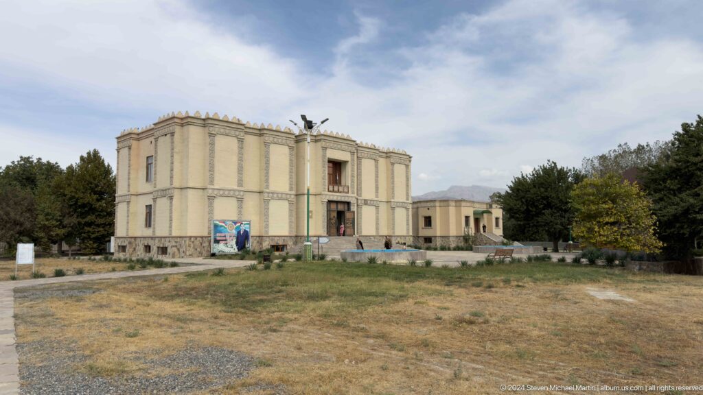 Distant view of the pale Ancient Sarazm museum complex across open dry ground under a cloudy sky with mountains behind.