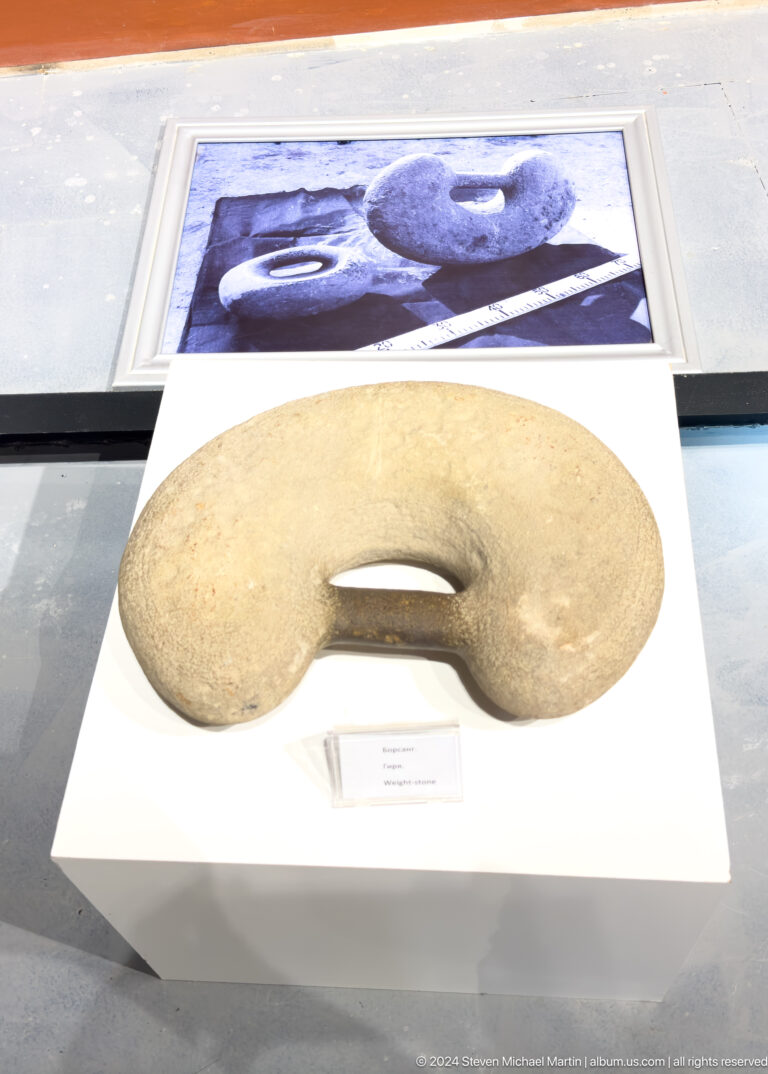 Broad crescent-shaped stone artifact from Sarazm with a rectangular cut-out in the center, shown in a museum display case.