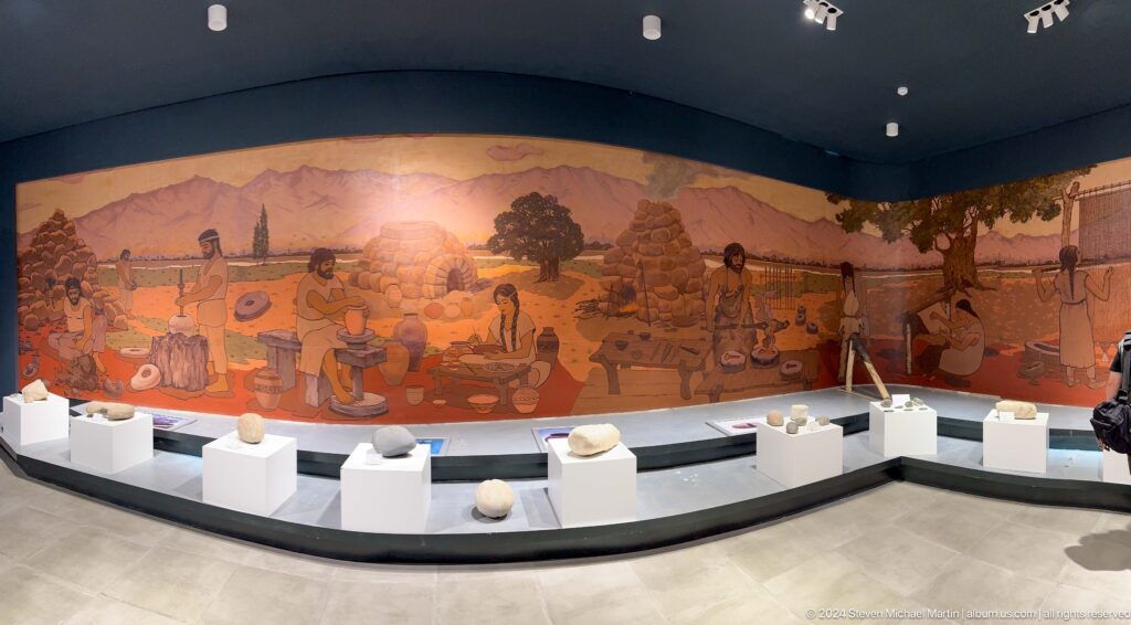 Panoramic mural of ancient Sarazm depicting people, animals, and huts above curved display cases of artifacts.