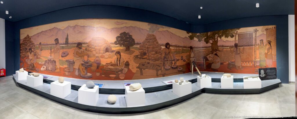 Curved mural reconstructing a prehistoric settlement in the Sarazm museum, with low white display cases of artifacts in the foreground.