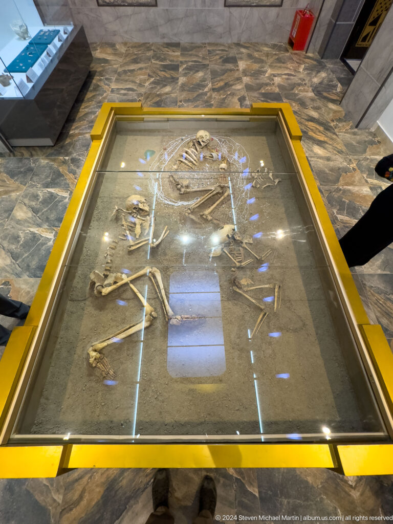 Museum display of an excavated Sarazm burial with a fully extended human skeleton and small artifacts arranged around it in a long glass case.