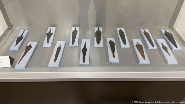 Row of dark stone and metal tools or weapons from Sarazm, each artifact mounted on a pale display backing.