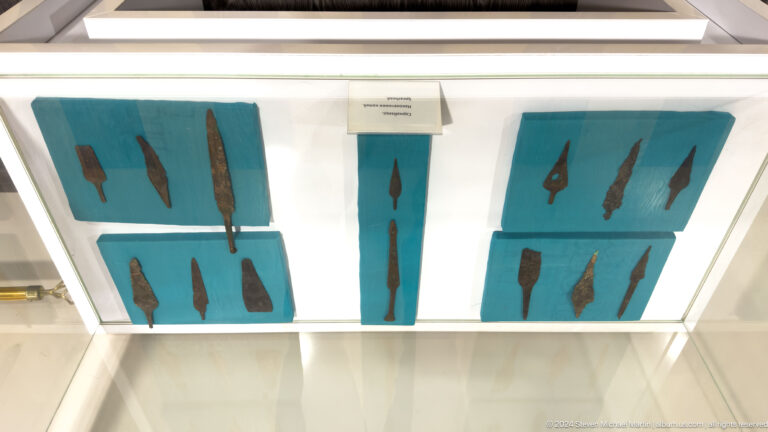 Angled view from below of a ceiling-mounted museum case displaying dark, elongated objects on turquoise panels from Sarazm.