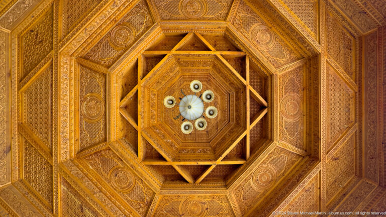 Octagonal carved wooden ceiling with dense geometric coffers and a central light in a traditional Tajik pavilion or museum space.