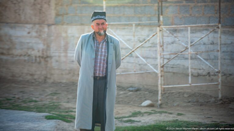 Elderly man in a traditional dark cap standing in a courtyard.