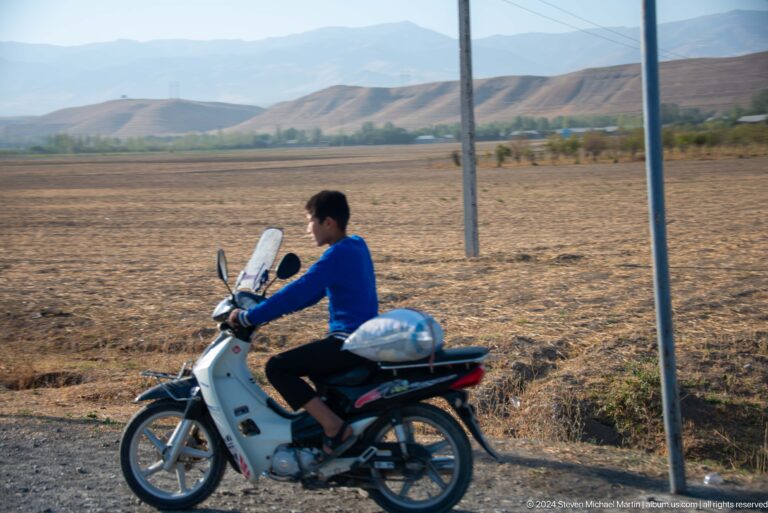 Man riding a white motor scooter through open farmland.