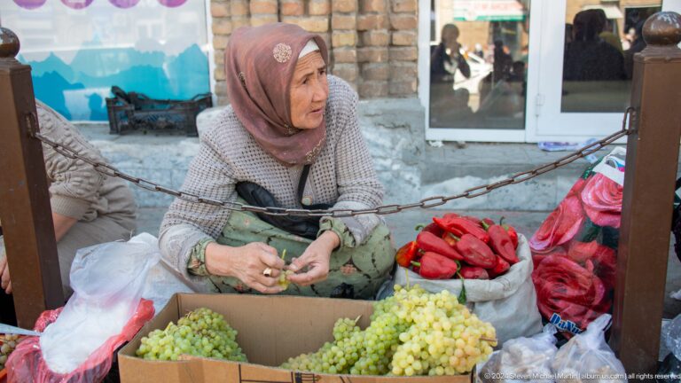 Woman vendor seated behind grapes and red peppers.
