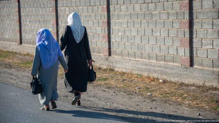 Two women in blue and black garments walking beside a block wall.