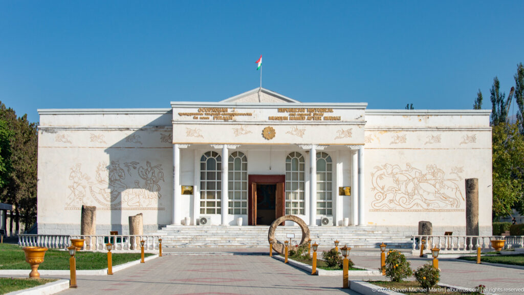 White museum facade with columned entrance, arched windows, Tajik flag, and linear wall murals.