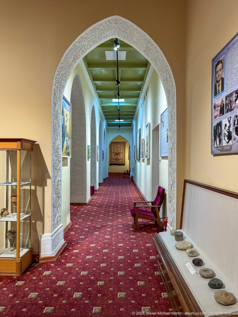 Long museum corridor with repeated white arches, red carpet, display cases, and wall exhibits.