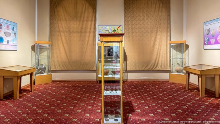 Carpeted museum room with three glass display cases containing pottery and small artifacts.