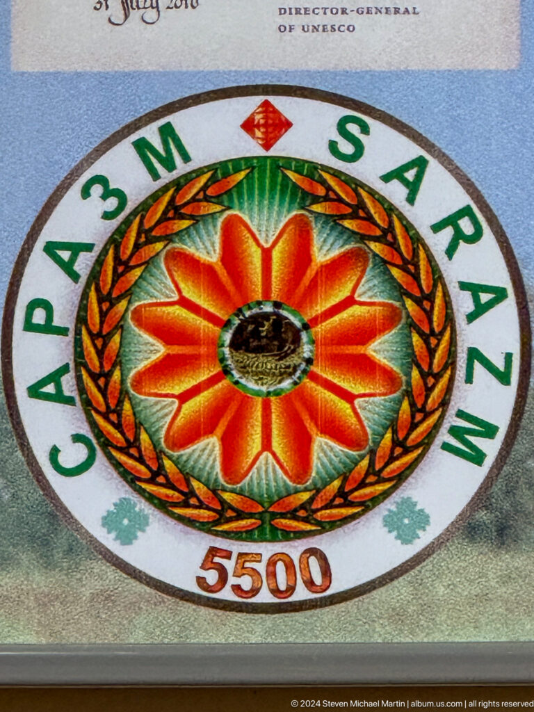 Circular SARAZM emblem with orange flower, central site image, and wreath-like border.