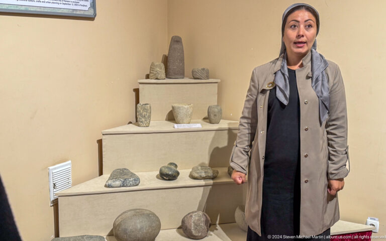 Museum staff member standing before tiered shelves of pottery, stone tools, and archaeological objects.