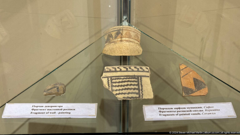 Glass case with painted ceramic fragments bearing black bands, zigzags, and triangular geometric motifs.