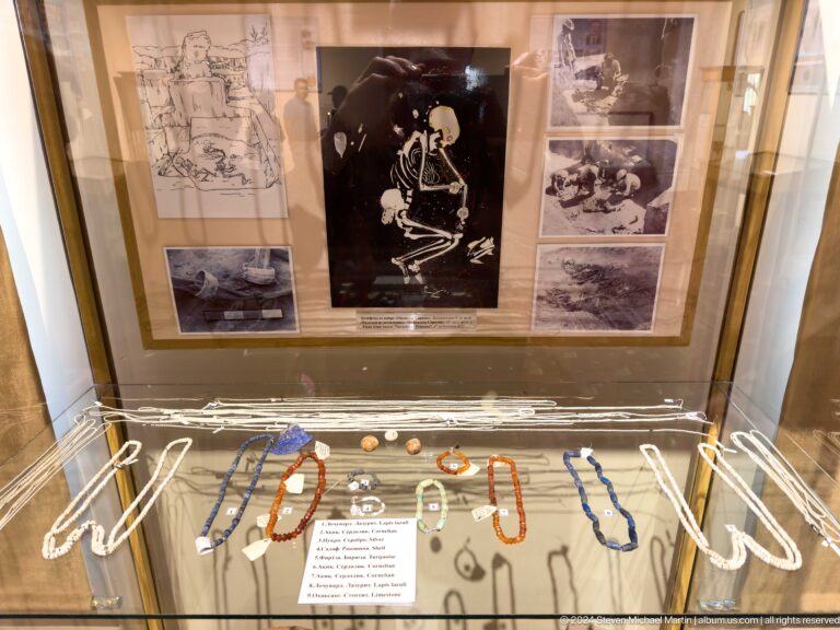 Glass case with colorful bead necklaces, excavation photographs, and a central skeletal reconstruction.